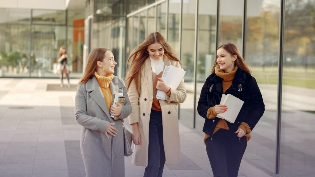 Kolme naista kävelee ulkona villakangastakeissa. Kaikki hymyilevät ja pitävät käsissään papereita ja kirjoja