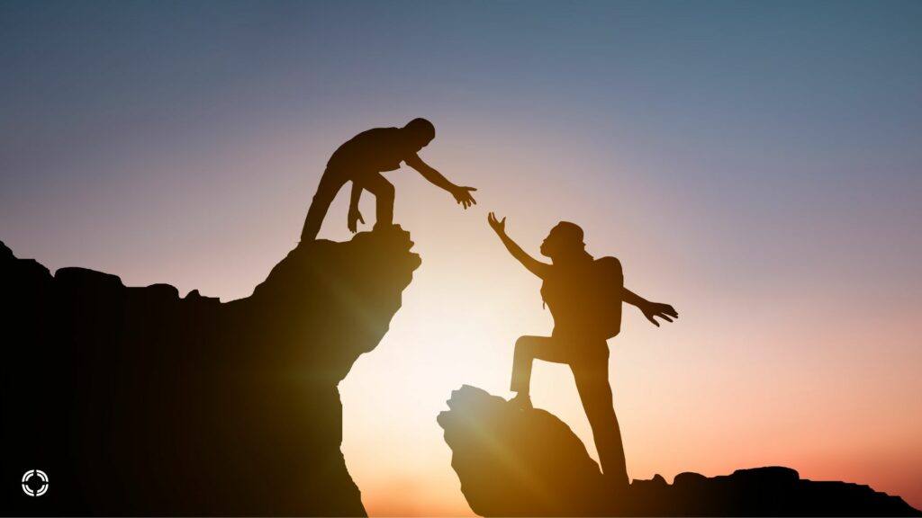 Kaksi ihmistä kurkottamalla toisiaan kohti auringonlaskun aikaan kalliolla.