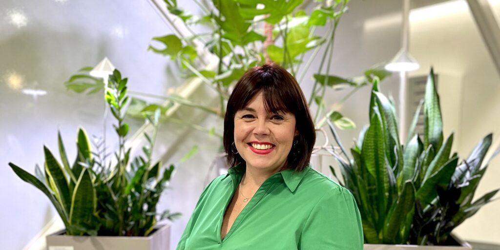 Minna Harju istumassa viherkasvien edessä Cursorin aulassa. Hänellä on päällään kirkkaan vihreä paita.